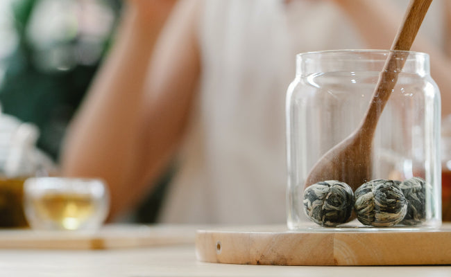 A glass jar with tea balls is pictured in the foreground, with a person next to a glass of tea making up the blurry background.