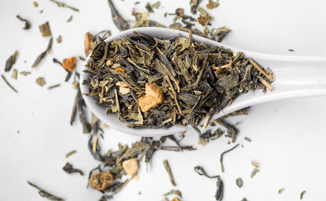 Top view of a white spoon with loose leaf tea ingredients. In the background, some of the ingredients can be seen having spilled onto the white surface below.