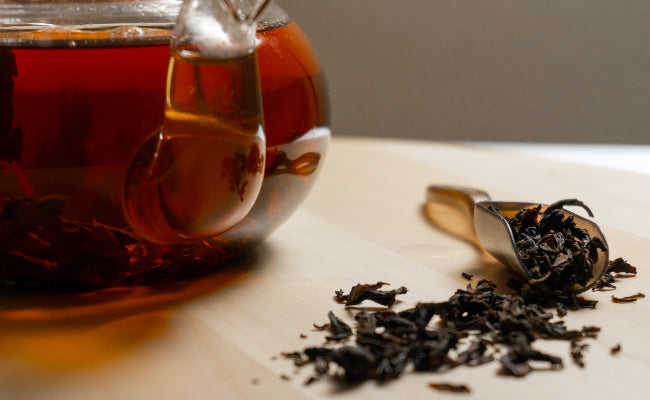 Close up of a glass teapot full of black tea, next to a tea scoop and some spilled black tea leaves.