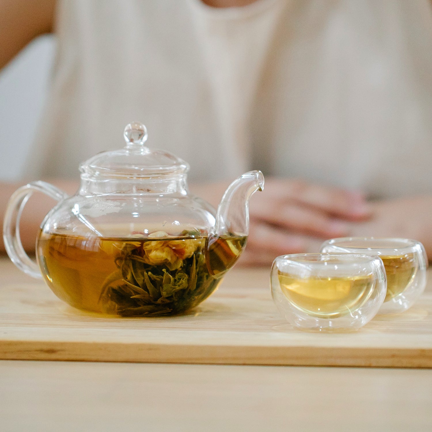 A glass teapot of loose leaf tea sits to the left of two glass cups of tea. In the blurred background, a person sites with their hands on on top of the other.