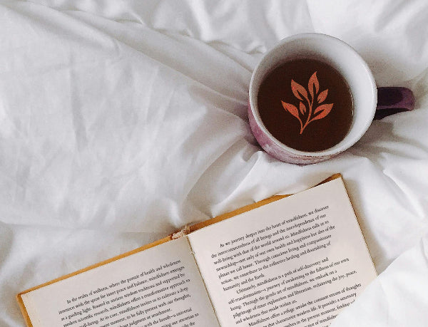 A book and a cup of tea are resting on a duvet, with the Botanika Tea logo visible in the tea.